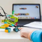 Dziecko buduje robota z klocków, ucząc się programowania. Komputer pokazuje interfejs do tworzenia kodu dla robotów w edukacji.