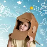 Dziecko w kartonowym hełmie astronauty patrzy w górę, otoczone rysunkami rakiet i planet. To właśnie takie marzenia o kosmosie napędzają nas do odkrywania.