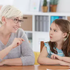 Kiedy z dzieckiem do logopedy: uśmiechnięta terapeutka z blond włosami i okularach pokazuje coś małej dziewczynce z kitkami.