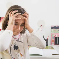 Mała dziewczynka z rękami na głowie, wyrażająca stres u dziecka w wieku przedszkolnym. Na biurku leżą zeszyty i kredki.