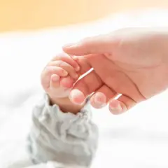 Dłoń dorosłego delikatnie obejmuje maleńką dłoń dziecka, ćwicząc chwyt pęsetkowy.
