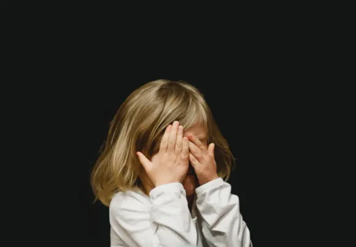 Mała dziewczynka z blond włosami zakrywa oczy dłońmi. Brak kontaktu wzrokowego, jakby grała w chowanego.