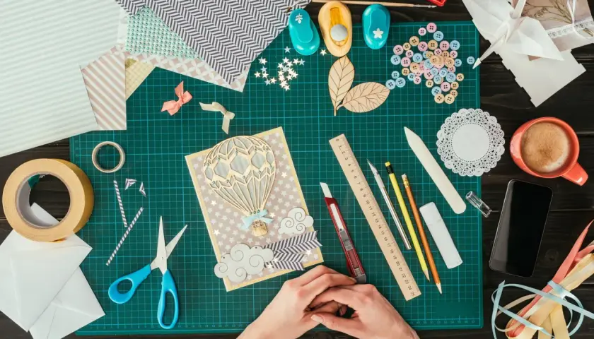 Ręce tworzą kartkę z balonem na gorące powietrze. Wokół materiały do scrapbookingu, taśma, nożyczki, guziki i inne akcesoria.