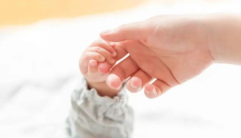 Dłoń dorosłego delikatnie obejmuje maleńką dłoń dziecka, ćwicząc chwyt pęsetkowy.