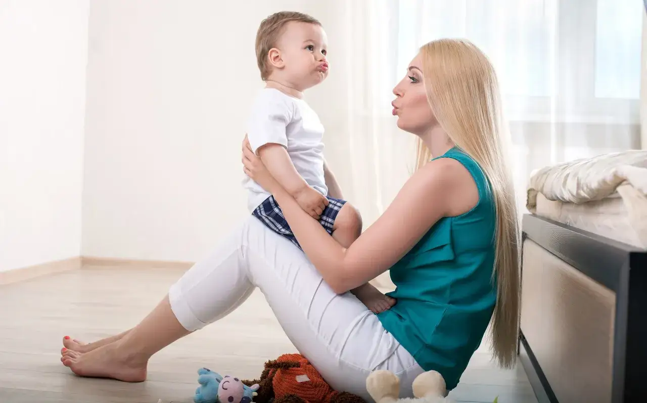 Mama uczy dziecko mówić, naśladując jego miny. W tle łóżko i zabawki.