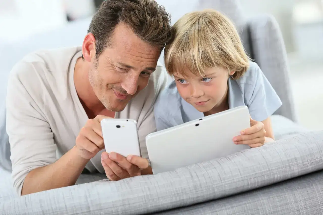Tata i syn leżą na kanapie, oglądając gry dla 5 latka na telefon i tablet.